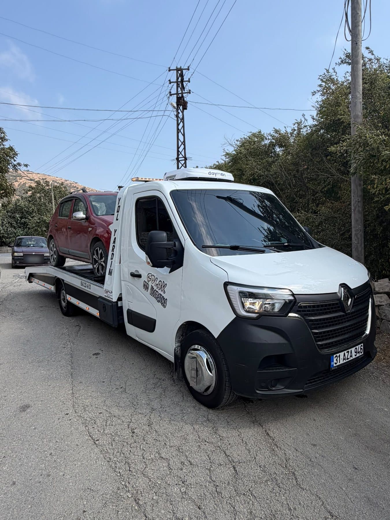 Mahalle arasında kasasında kırmızı hatchback taşıyan Çolak Oto Kurtarma çekicisi